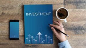 picture showing a phone, a coffee cup and a book with the word ‘investment’. A hand holding a pen getting ready to write in the investment book