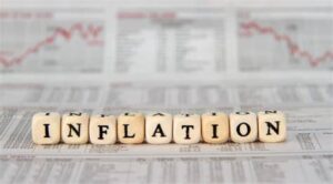 The word “Inflation” is written in wood letters shaped in dice on a background of papers that show statistics and graphs