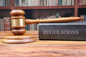 A wooden judge’s gavel rests on a round sound block beside a large book labelled “Regulations.”