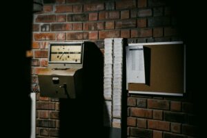 An old punch clock on a brick wall, next to a rack of time cards and a cork board with a sheet of paper pinned to it